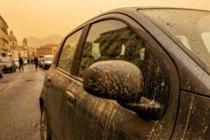 Primo piano di uno specchietto laterale di un'auto scura coperta da fango e polvere, con goccioline di pioggia visibili sulla superficie. L'auto è parcheggiata su una strada a Napoli, dove gli edifici e le cupole della città si intravedono sullo sfondo sotto un cielo cupo e nuvoloso.