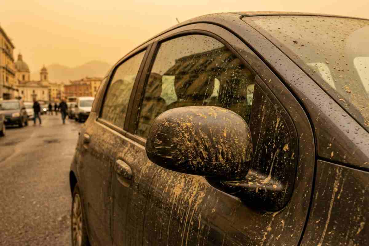 Primo piano di uno specchietto laterale di un'auto scura coperta da fango e polvere, con goccioline di pioggia visibili sulla superficie. L'auto è parcheggiata su una strada a Napoli, dove gli edifici e le cupole della città si intravedono sullo sfondo sotto un cielo cupo e nuvoloso.